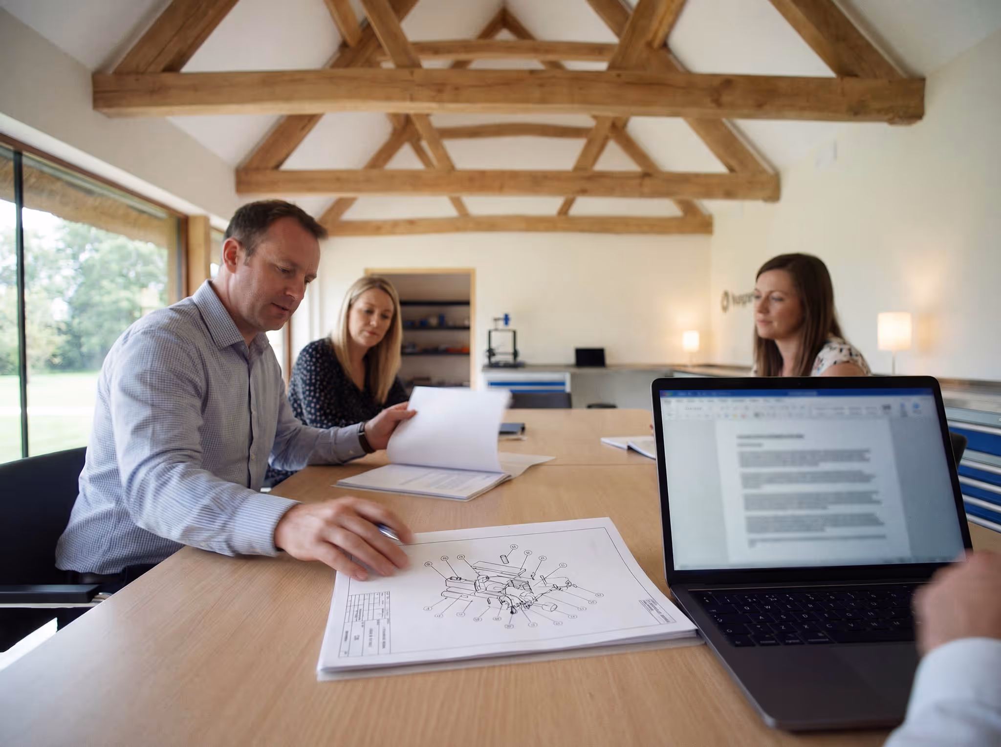 Three people in a meeting room with wooden beams on the ceiling, reviewing technical documents and a laptop displaying a text document.