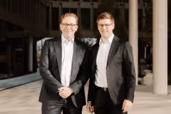 Rechtsanwalt Marius Siemen und Steuerberater Lukas Conrady in schwarzen Anzügen und weißen Hemden stehen nebeneinander im Freien vor einem modernen Gebäude.
