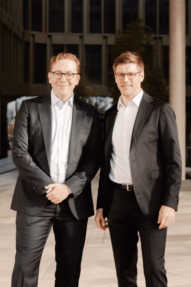 Zwei Männer in schwarzen Anzügen mit weißen Hemden und Brillen stehen nebeneinander vor einem modernen Gebäude.