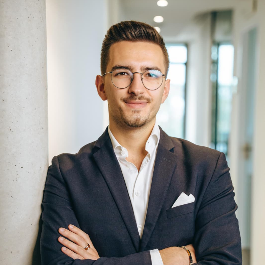 Ein junger Mann mit Brille, weißem Hemd und dunklem Sakko lehnt mit verschränkten Armen an einer Säule in einem modernen Büroflur.
