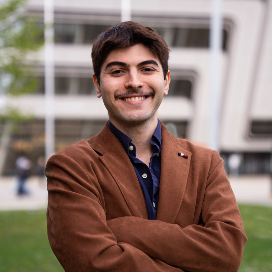 Junger Mann mit braunem Sakko und dunklem Hemd, lächelnd mit verschränkten Armen im Freien vor einem modernen Gebäude.