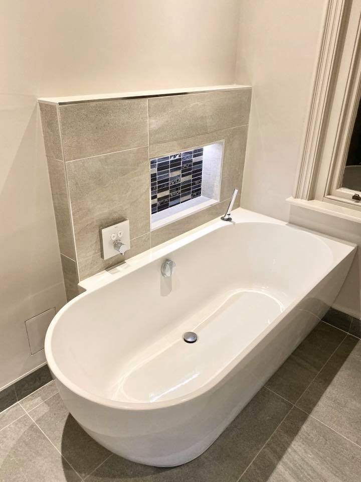Modern white freestanding bathtub with tiled surround and built-in niche in a clean bathroom.