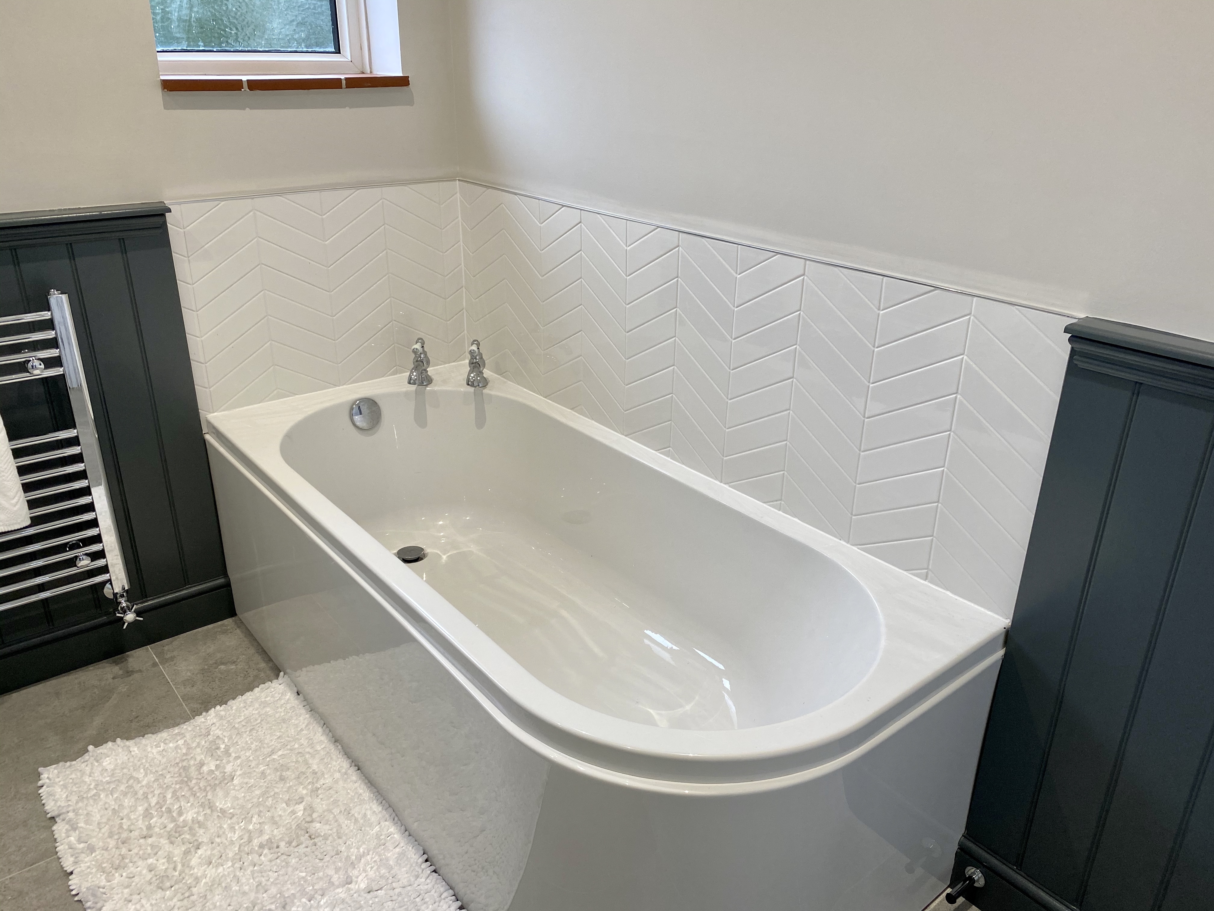 White bathtub with silver faucets in a bathroom with white chevron tiles and dark wainscoting.