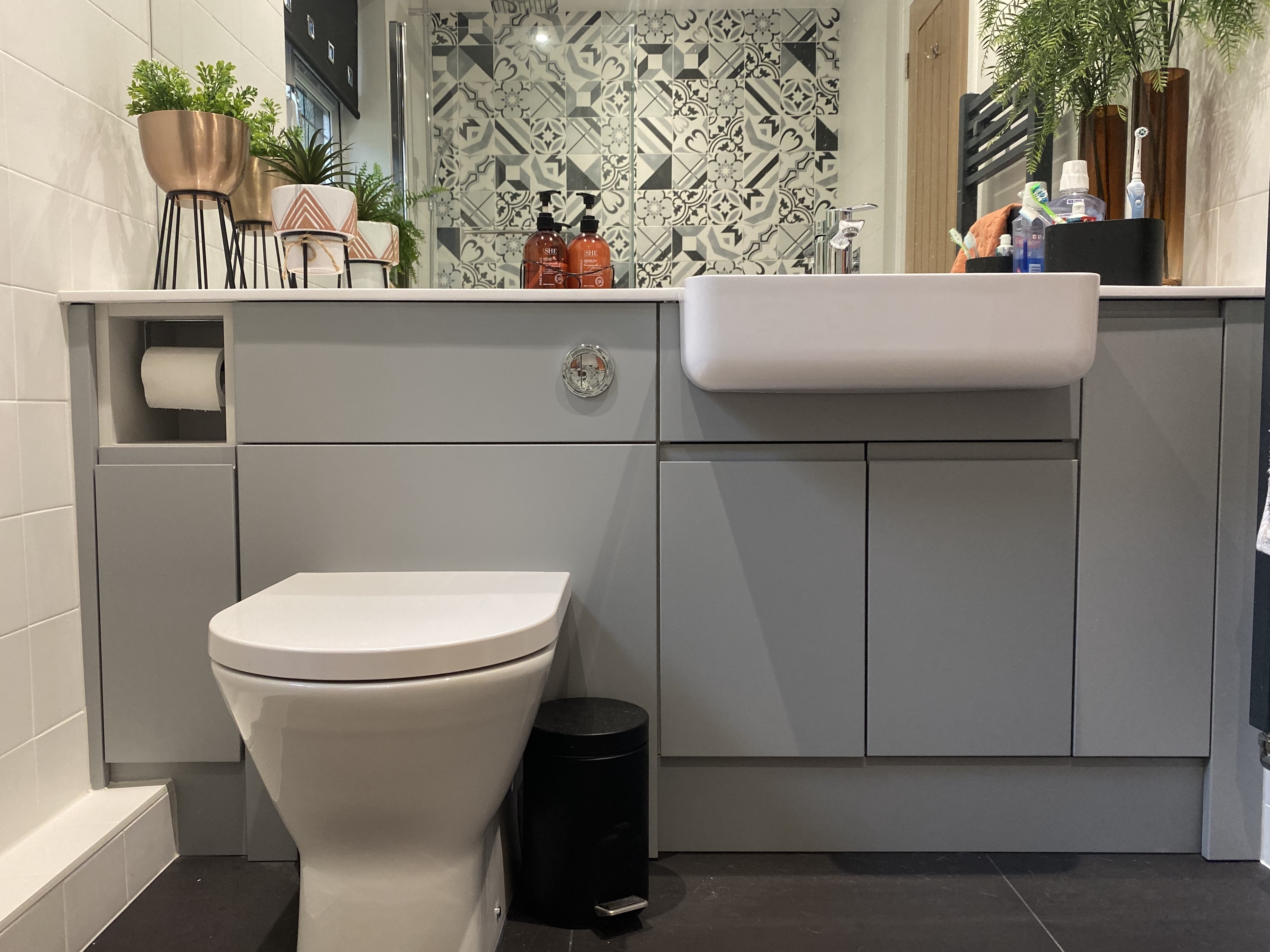 Modern bathroom with white toilet, gray cabinets, white sink, plants, and patterned tile wall.