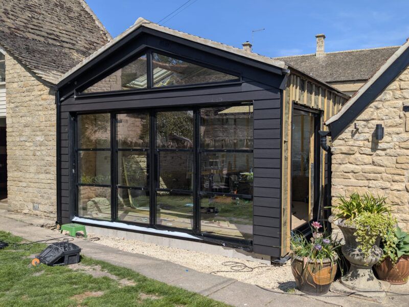 New black-framed glass extension on rustic stone house with visible interior garden reflection.