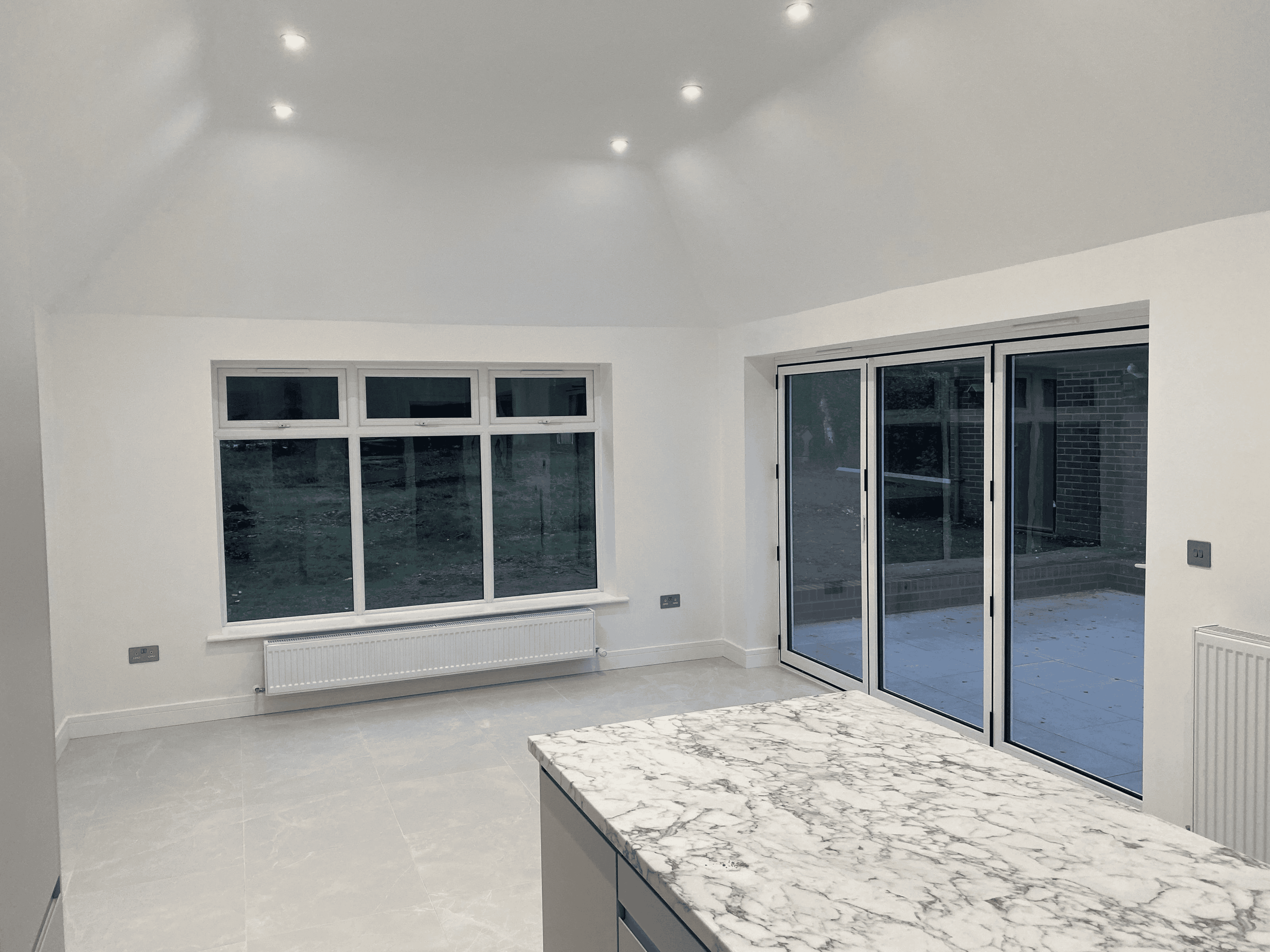 Empty modern kitchen with marble island, large windows, sliding glass doors, white walls, and ceiling lights.