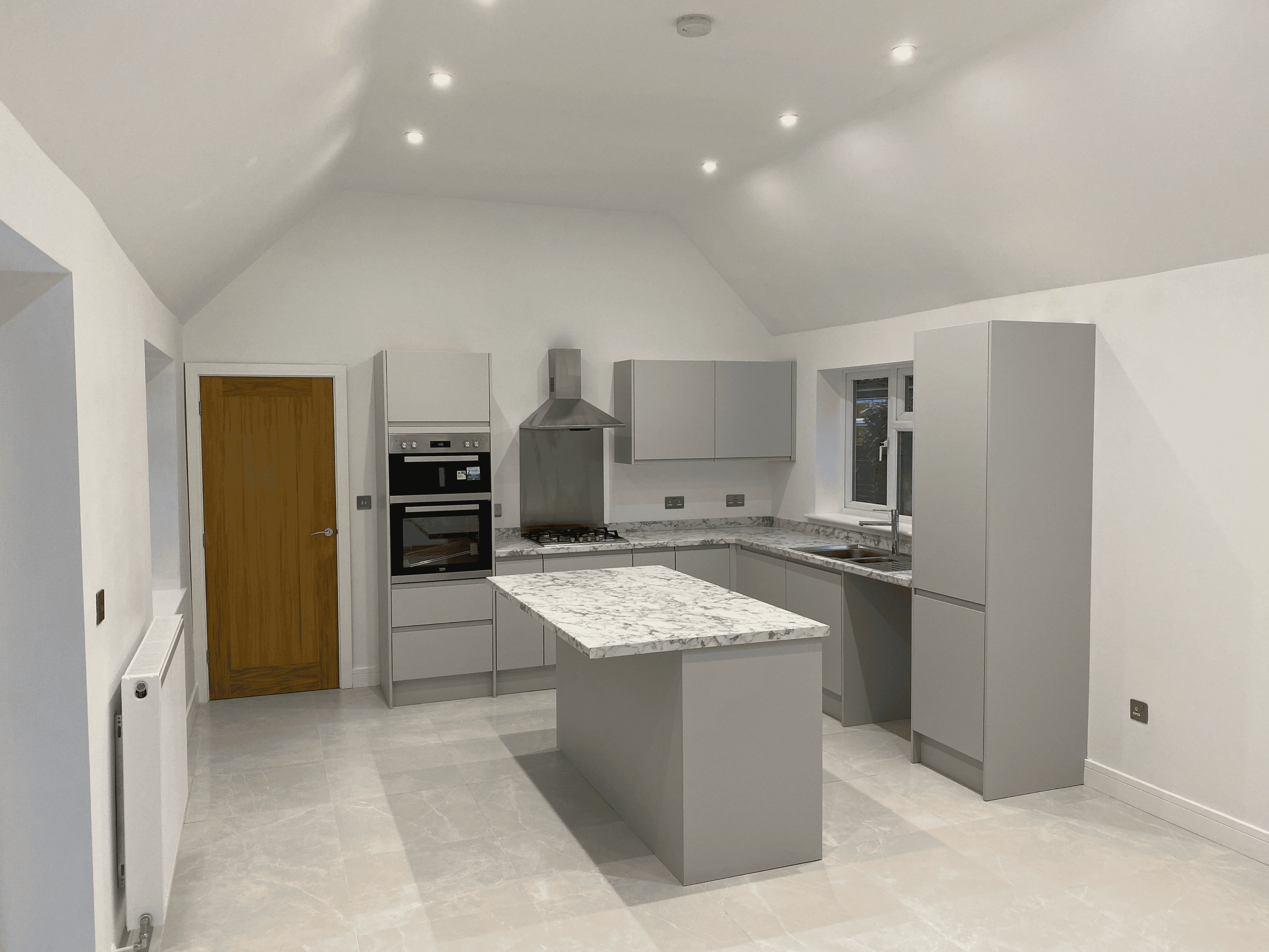 Modern kitchen with gray cabinets, marble countertops and island, wooden door, and recessed ceiling lights.