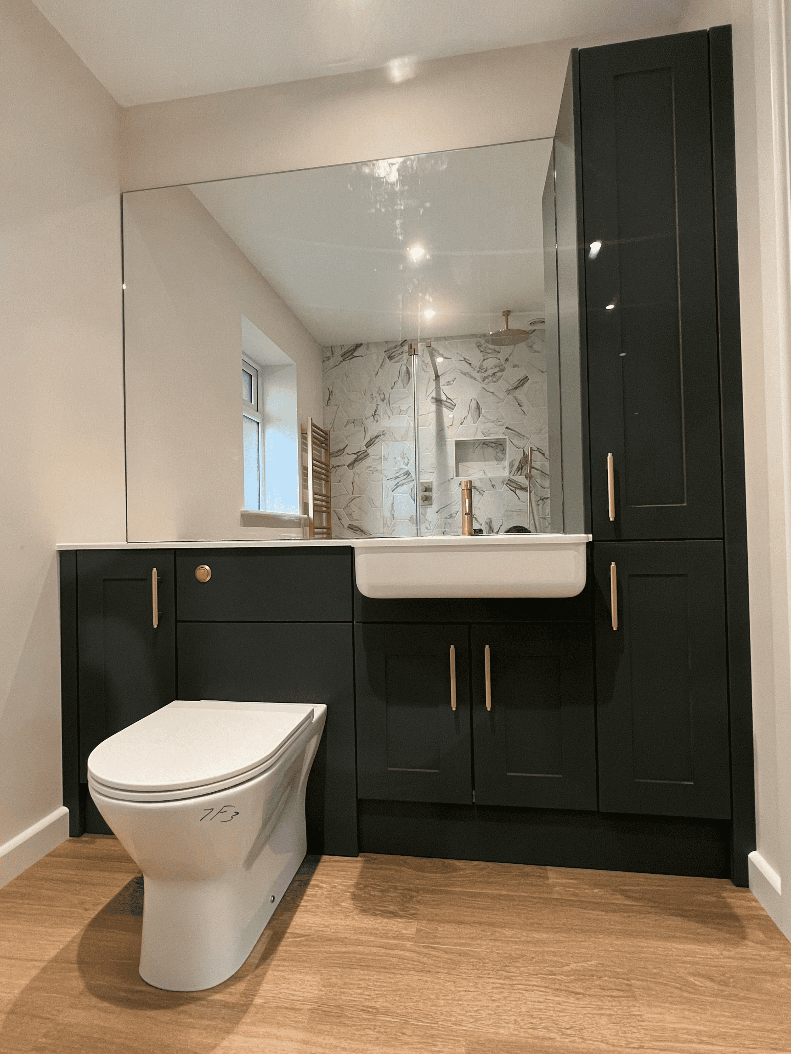 Modern bathroom with wooden floor, dark cabinetry, white sink, toilet, and large mirror reflecting marble shower.