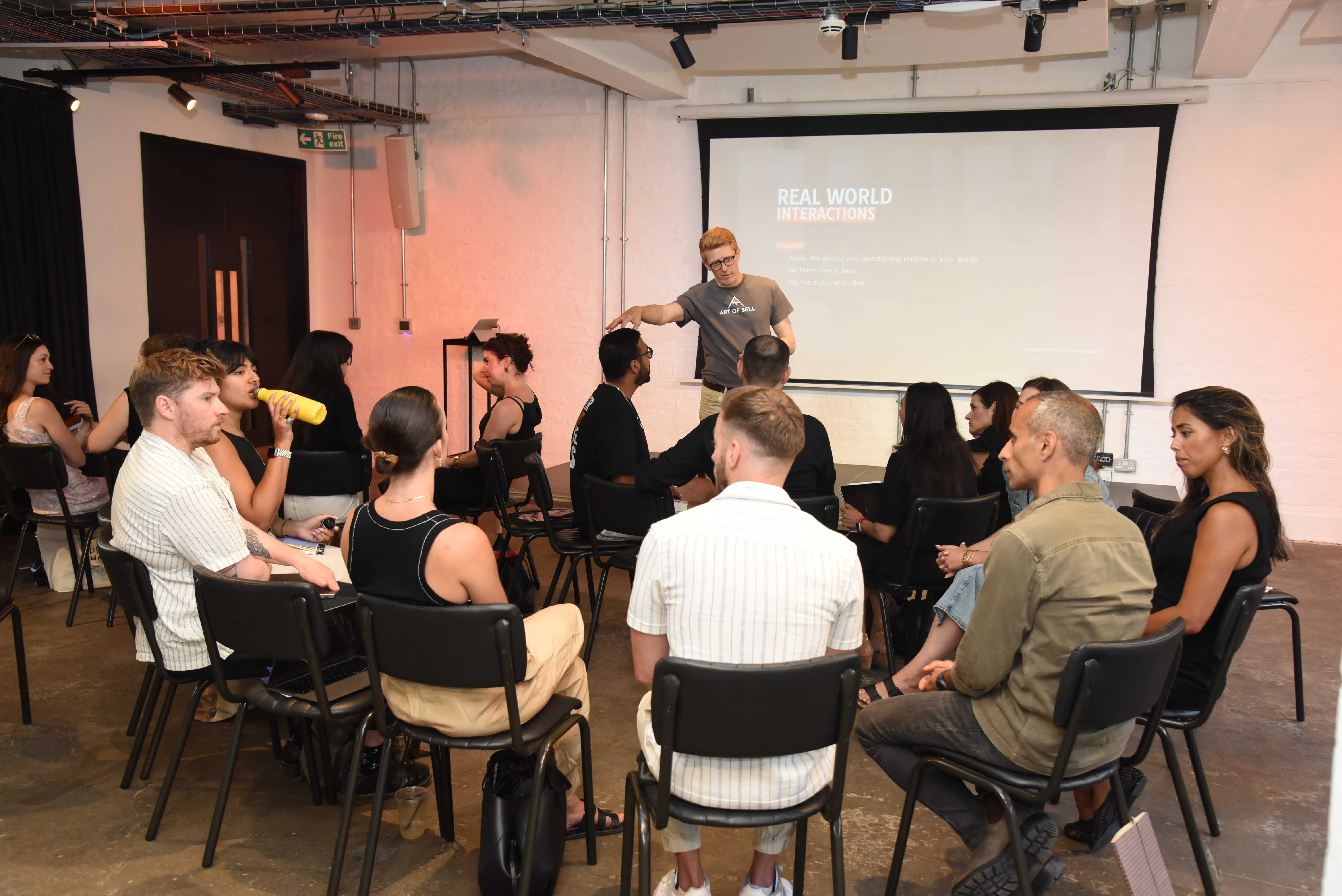 Group in a workshop room with speaker discussing in front of a presentation screen titled Real World Interactions.