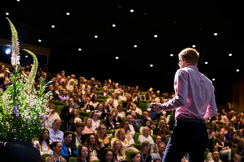 Man in pink shirt speaking to a large audience in a dark auditorium with green seats.