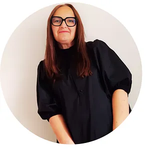 Woman with long brown hair and glasses wearing a black dress against a plain light background.