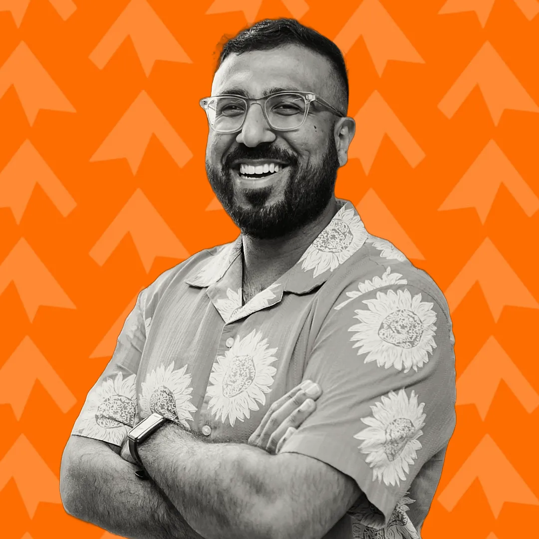 Smiling man with glasses and beard, wearing a sunflower shirt, arms crossed on orange patterned background.