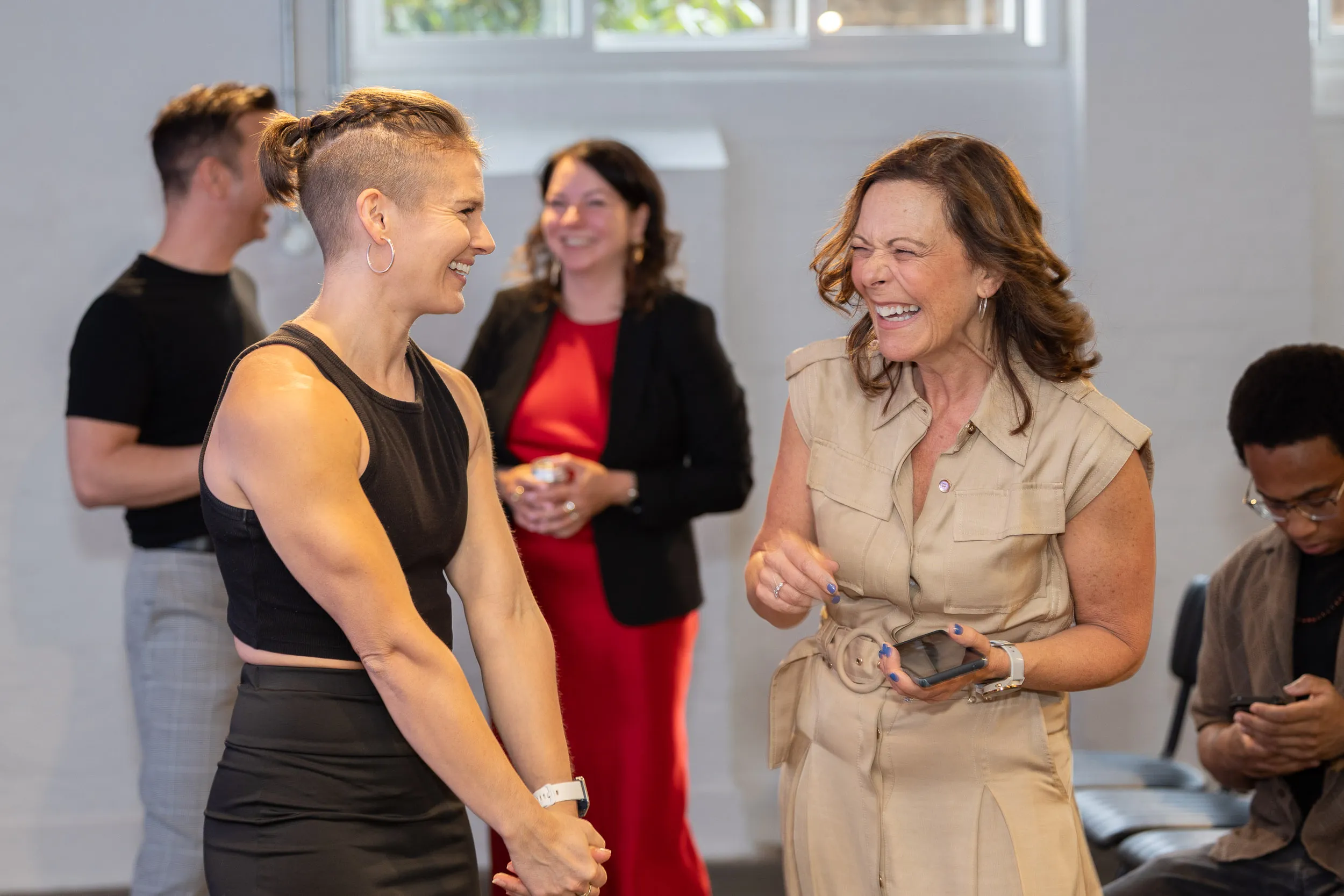 Two women smiling and laughing while talking in a bright indoor setting with three people in the background.