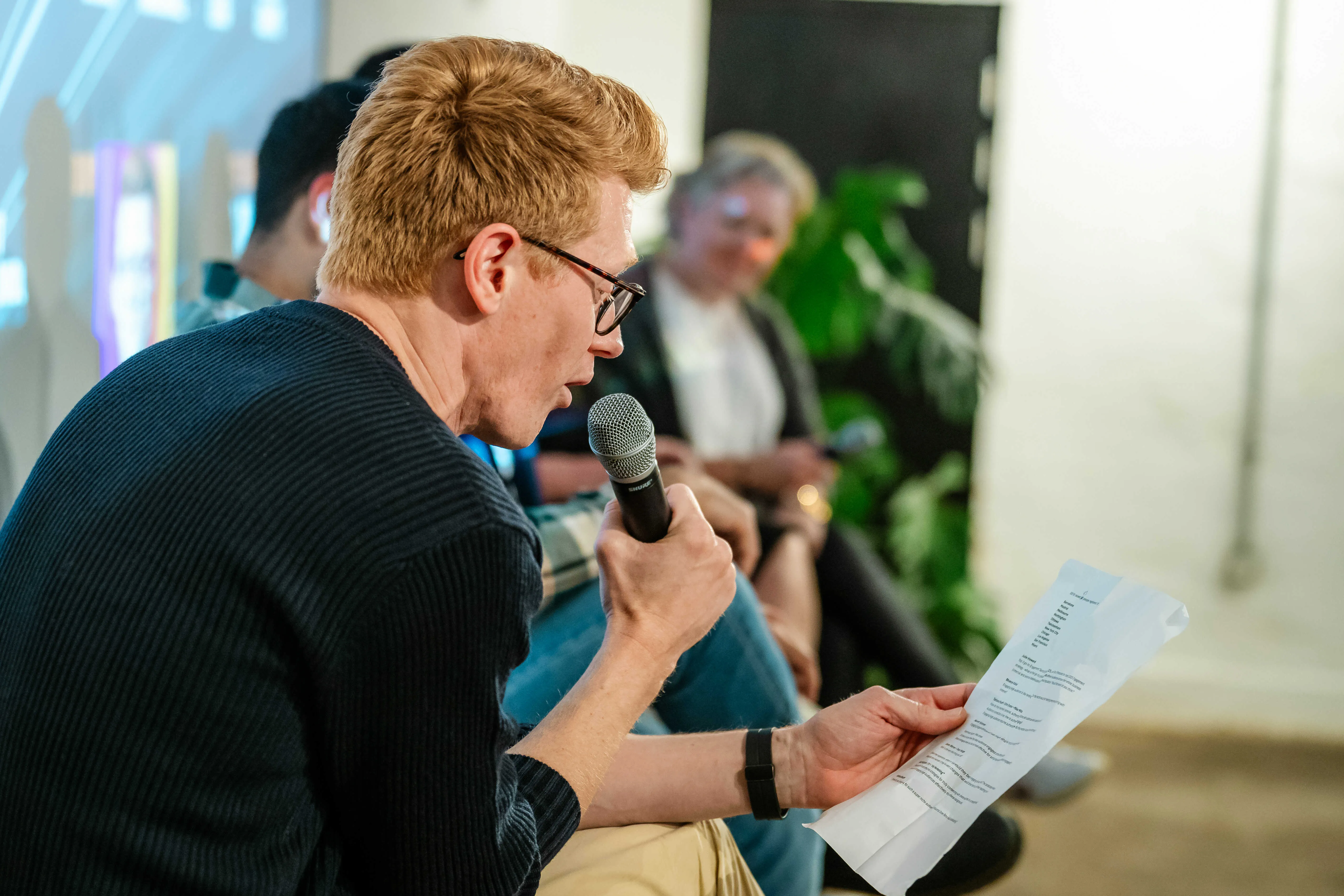 A man with glasses holds a microphone and reads from a paper during an indoor discussion event.