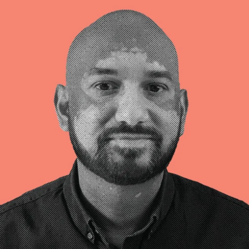 Portrait of a bald man with a beard wearing a buttoned shirt against a coral background.