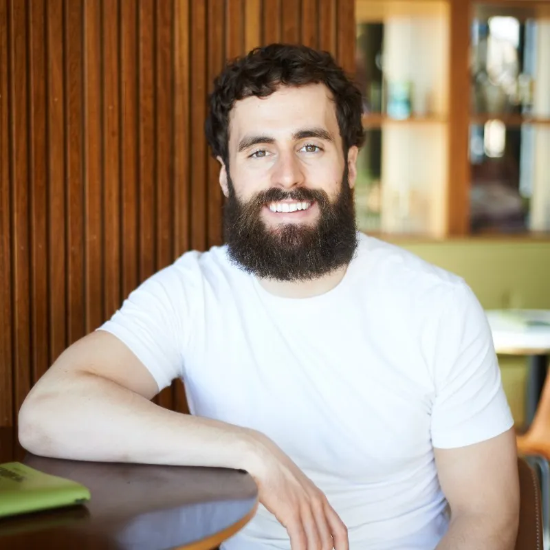 Bearded man in a white shirt smiling, sitting at a round table with a wooden panel background.