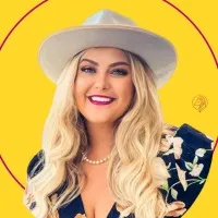 Smiling woman with long blonde hair wearing a white hat, pearl necklace, and floral dress against yellow background.