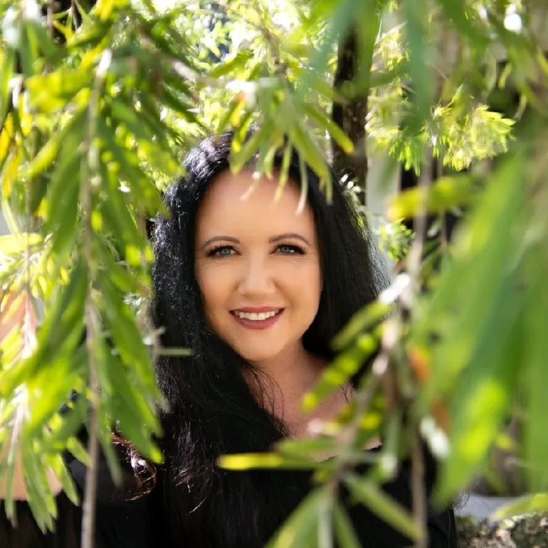 Smiling woman with long black hair framed by green foliage in natural outdoor light.