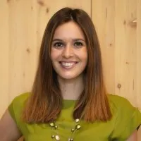 Smiling woman with straight brown hair wearing a green top and a beaded necklace, in front of wooden background.