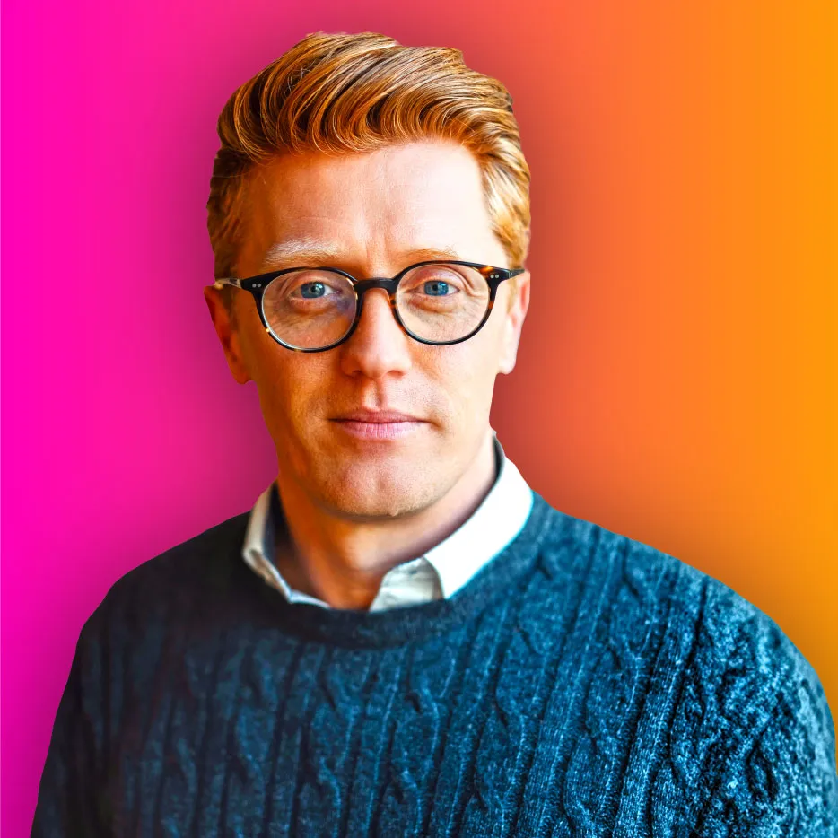 Man with red hair and glasses wearing a blue sweater against a pink and orange gradient background.