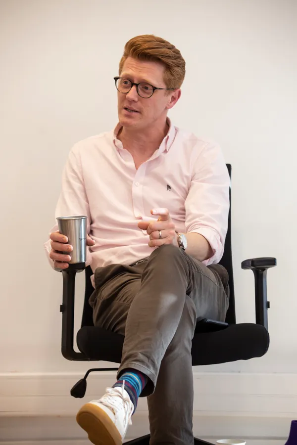 Man with glasses in a pink shirt sitting in an office chair holding a metal cup and gesturing.