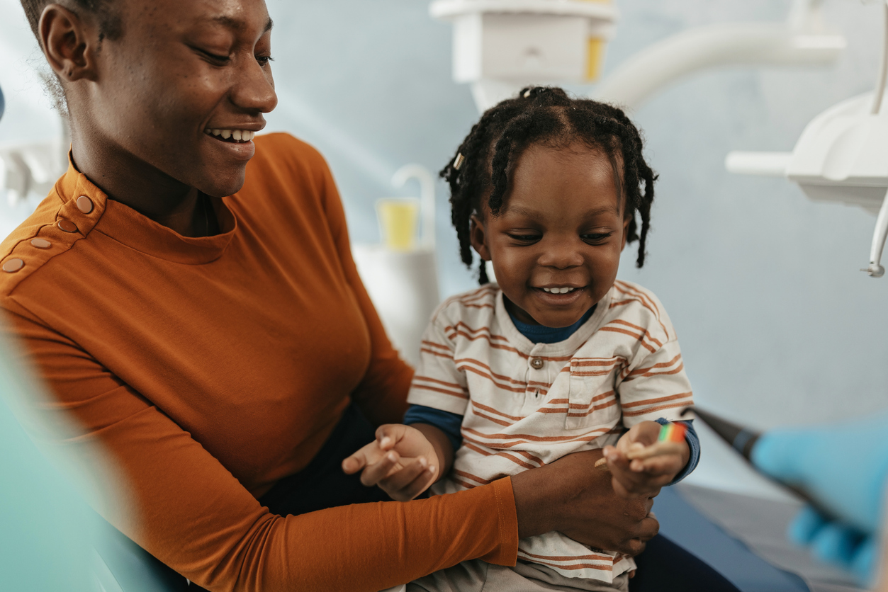Smiling mother holding her young son in a dental clinic while a gloved hand shows a colorful dental tool.