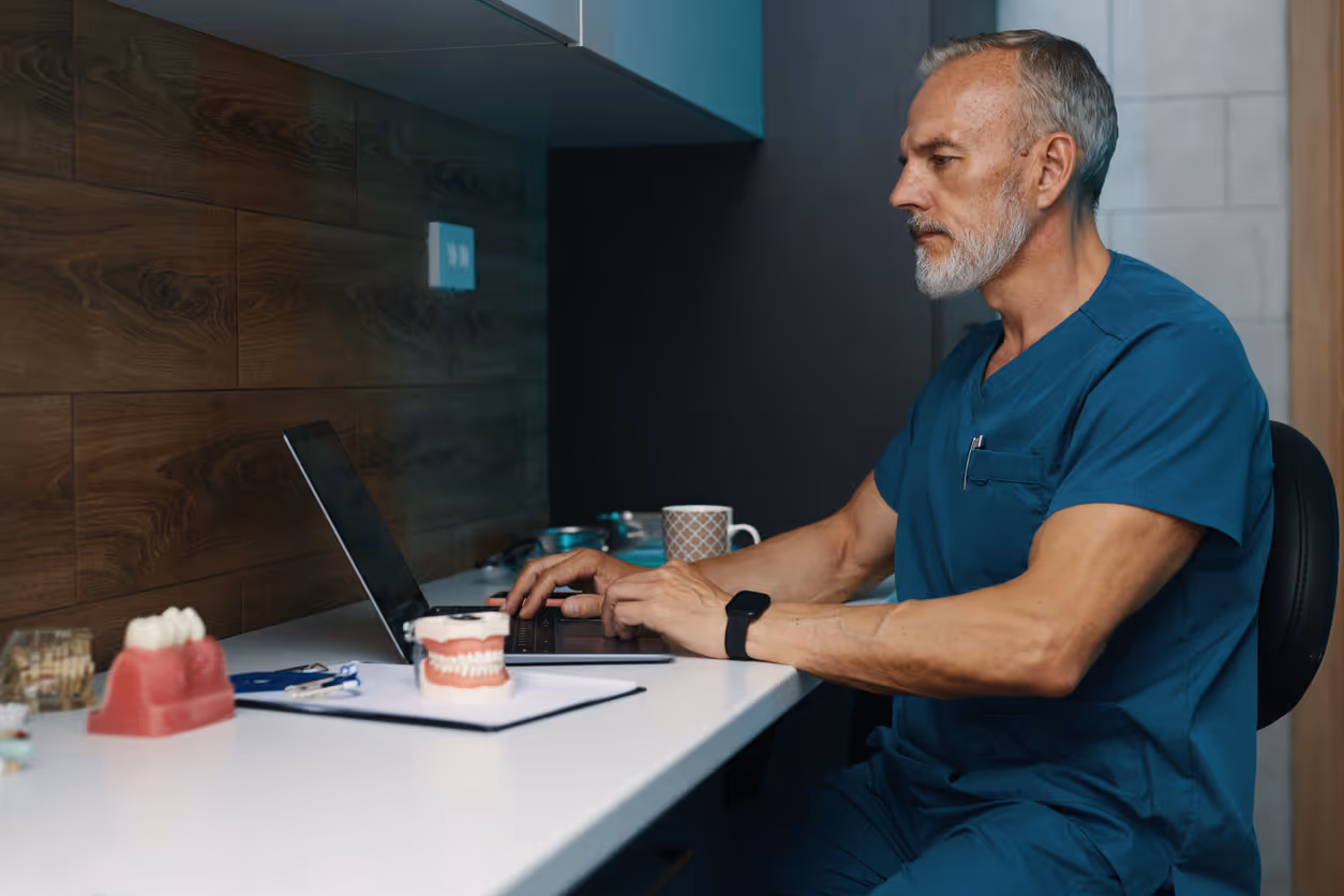 Male dentist in blue scrubs working on a laptop at a white desk with dental models and tools.