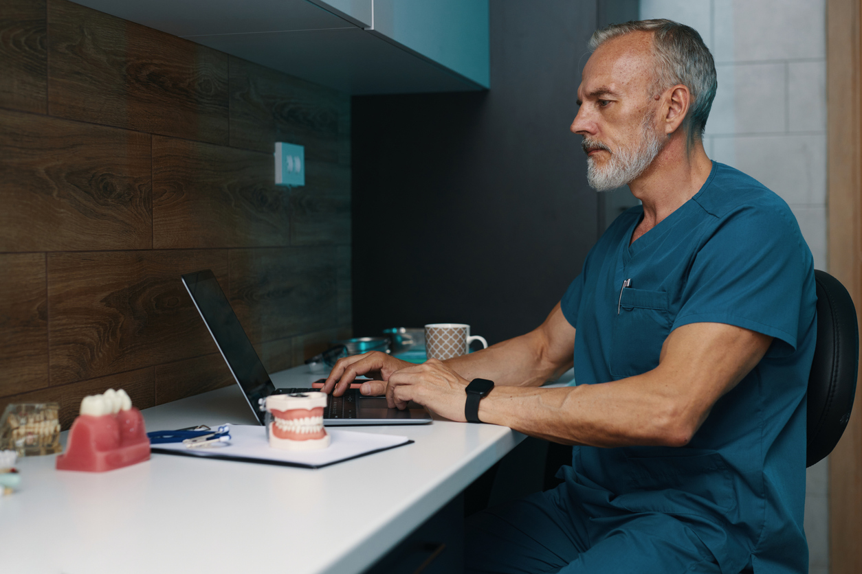 Male dentist in blue scrubs working on a laptop at a white desk with dental models and tools.