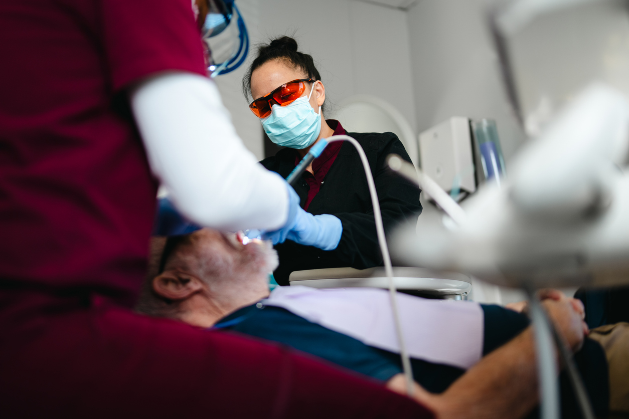 Dental professionals wearing masks and gloves examining an elderly male patient's teeth during a dental checkup.