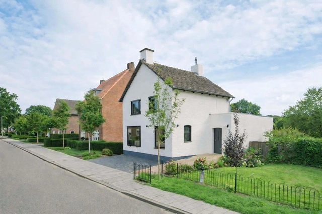 White two-story house with a brown shingled roof, small front yard with trees and plants, next to a sidewalk and street in a suburban neighborhood.