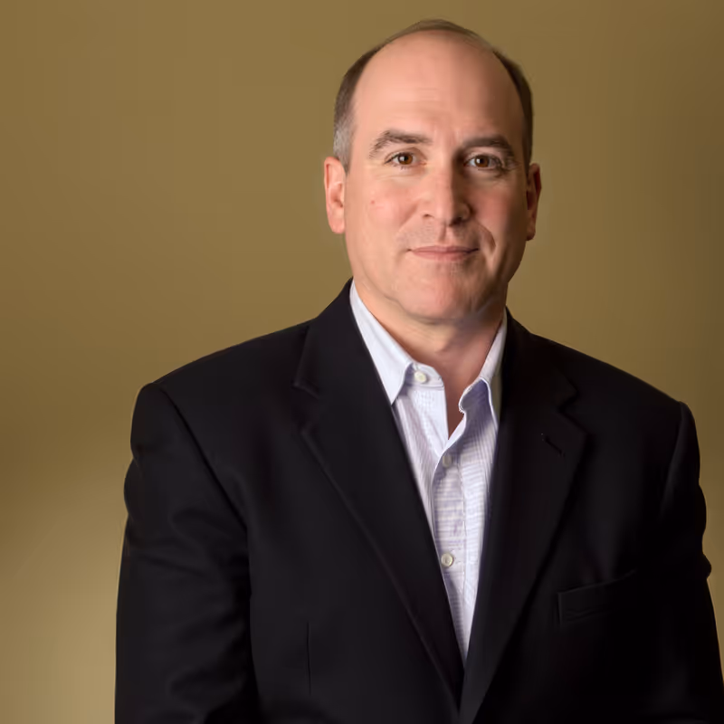 Portrait of a middle-aged man wearing a black suit jacket and light blue shirt against a plain brown background.