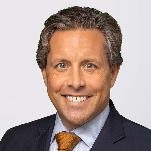 Smiling middle-aged man with gray hair wearing a dark suit, light blue shirt, and brown tie against a plain background.