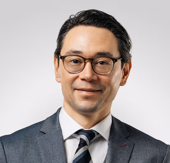 Professional man with black hair and glasses wearing a dark suit, white shirt, and striped tie against a light gray background.