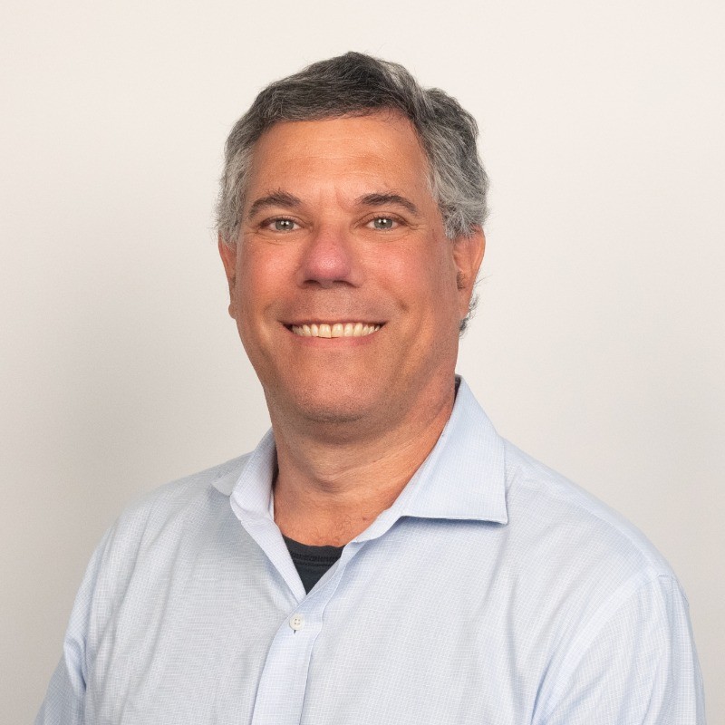 Smiling middle-aged man with gray hair wearing a light blue button-up shirt against a plain background.