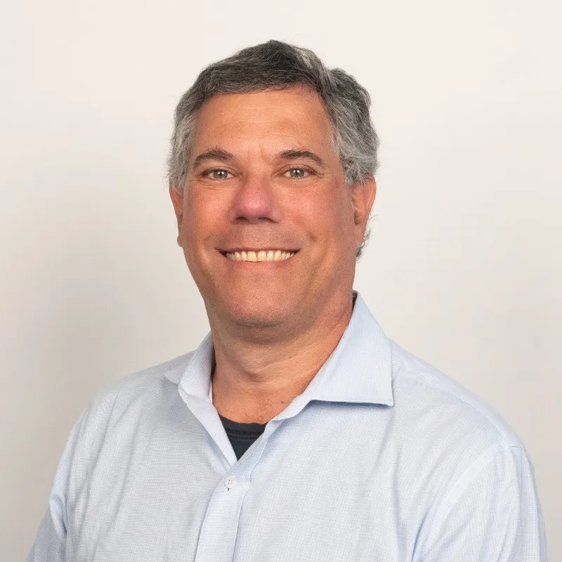 Smiling middle-aged man with gray hair wearing a light blue button-up shirt against a plain background.
