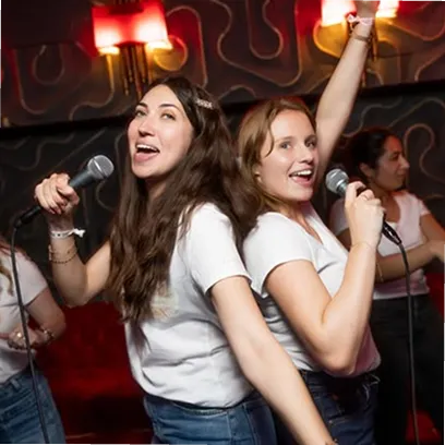 Two young women back-to-back singing energetically into microphones in a dimly lit karaoke setting.