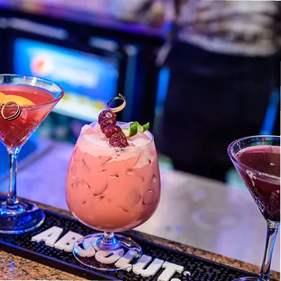 Three colorful cocktails on a bar counter with one pink drink garnished with cherries and lime wedge in the center.