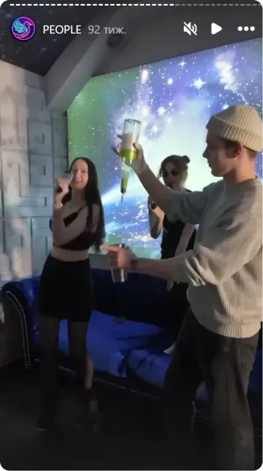 Three young adults enjoying drinks in a dimly lit room with a colorful cosmic background projection.