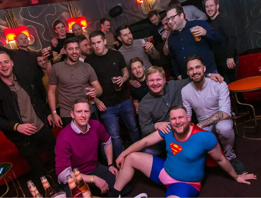 Four friends enjoying a karaoke party, two women wearing star-shaped glasses and holding microphones, one wearing a birthday crown, with a pink 'Happy Birthday' hat on a drum table.