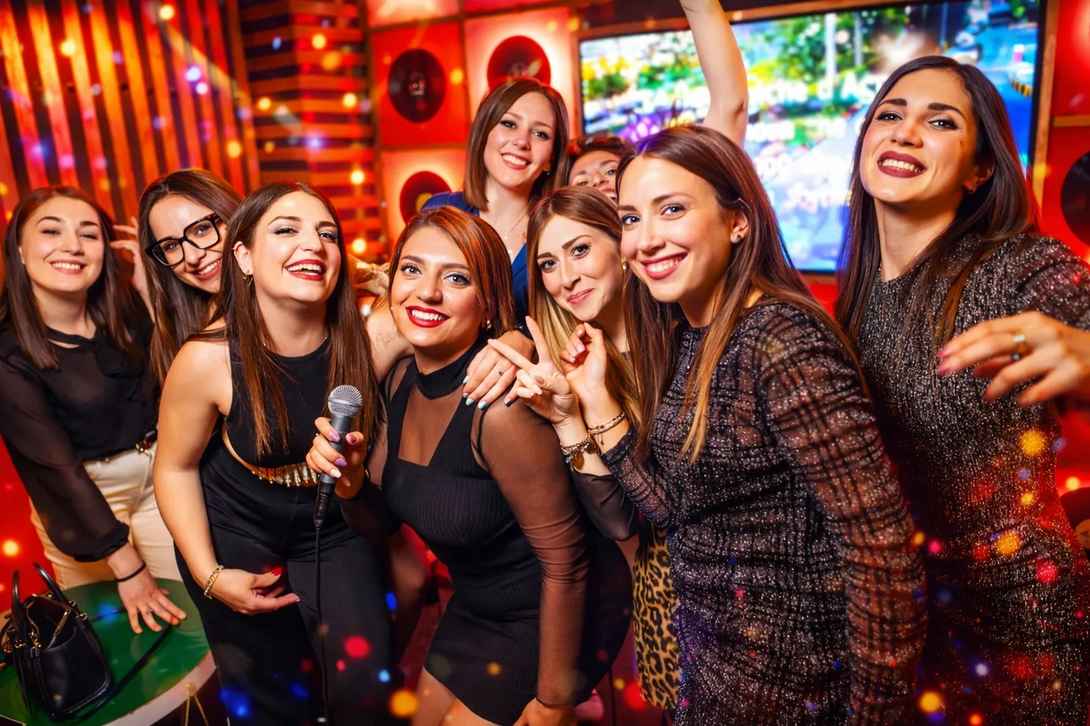 Four friends enjoying a karaoke party, two women wearing star-shaped glasses and holding microphones, one wearing a birthday crown, with a pink 'Happy Birthday' hat on a drum table.