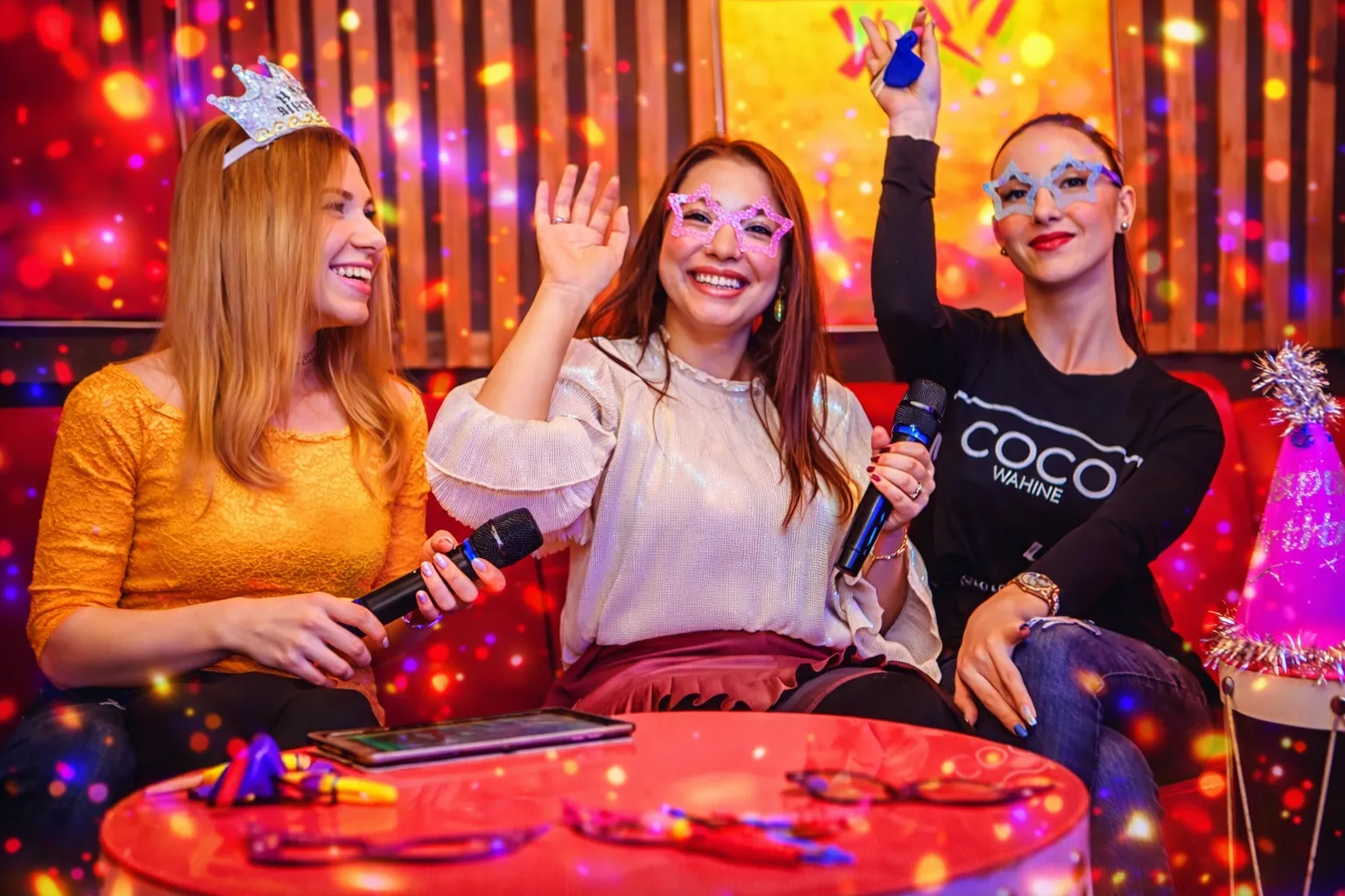 Four friends enjoying a karaoke party, two women wearing star-shaped glasses and holding microphones, one wearing a birthday crown, with a pink 'Happy Birthday' hat on a drum table.