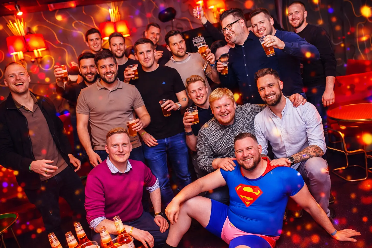 Four friends enjoying a karaoke party, two women wearing star-shaped glasses and holding microphones, one wearing a birthday crown, with a pink 'Happy Birthday' hat on a drum table.