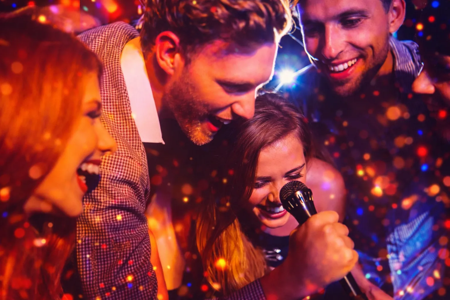 Four friends enjoying a karaoke party, two women wearing star-shaped glasses and holding microphones, one wearing a birthday crown, with a pink 'Happy Birthday' hat on a drum table.