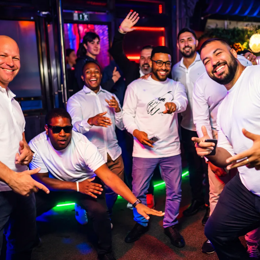 Group of eight men smiling and posing energetically at a nighttime indoor event with colorful lighting.