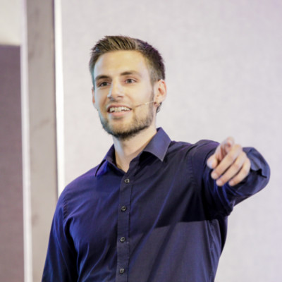 Man with a beard and short hair wearing a dark blue shirt speaking with a microphone headset and pointing forward.