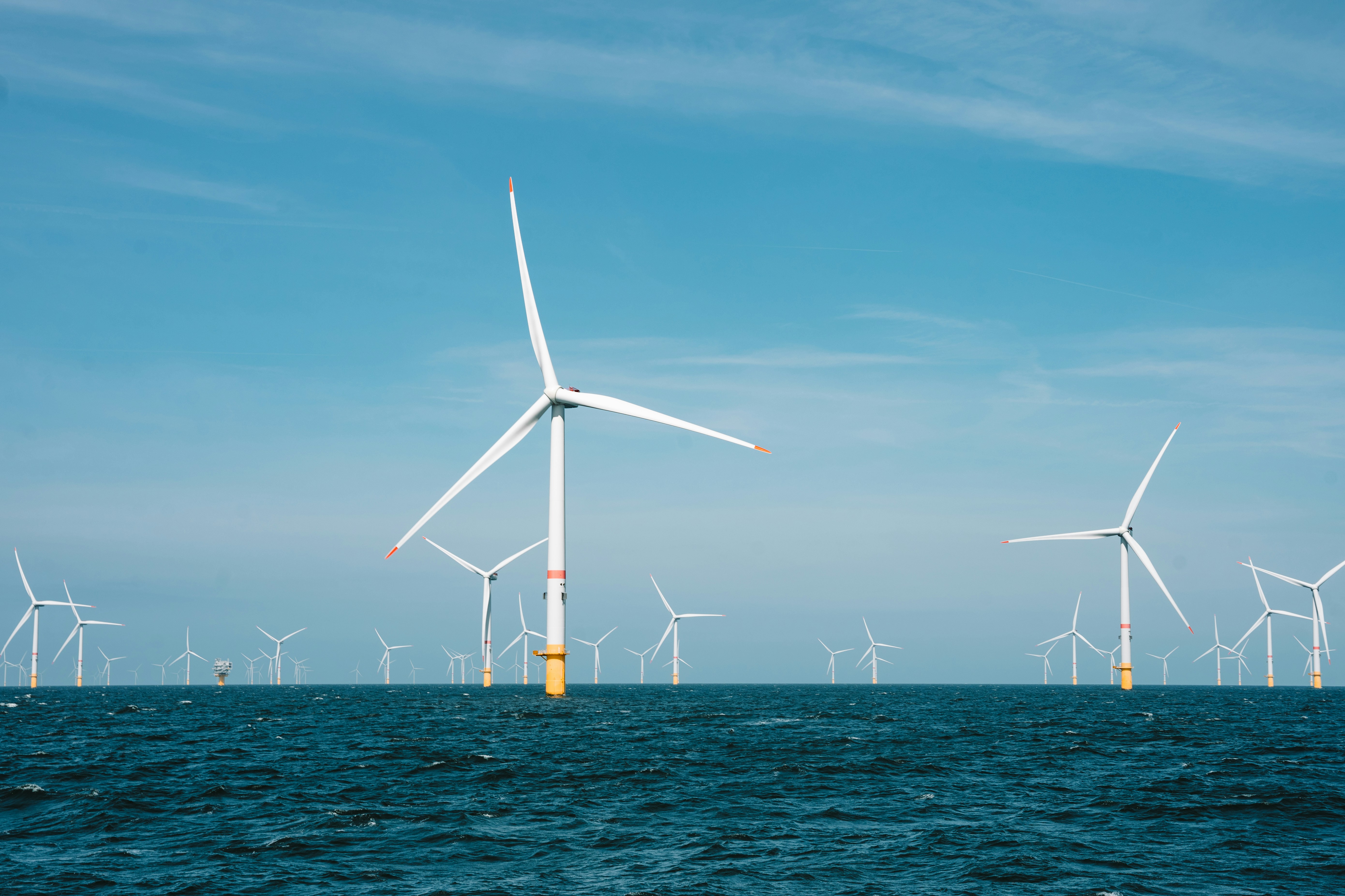 Offshore wind farm with multiple large white wind turbines in a blue ocean under a clear sky.