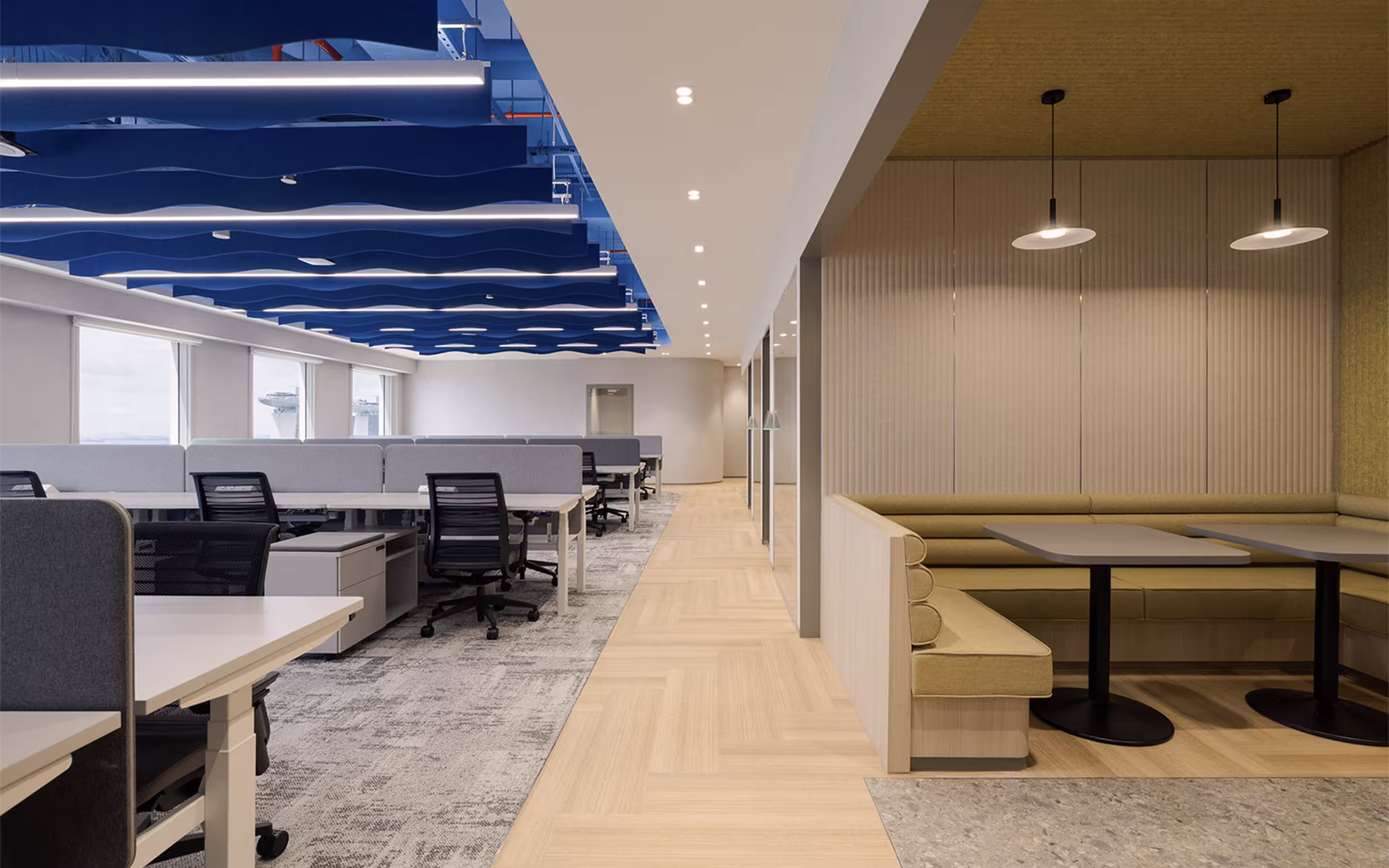 Modern office space with rows of desks and black swivel chairs under blue wave-like ceiling panels, and a cozy seating booth with pendant lights on the right.