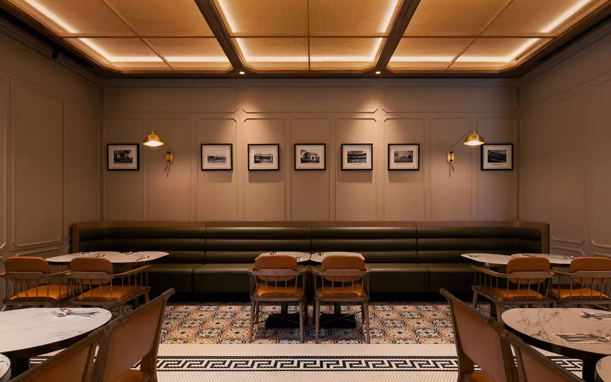 Elegant restaurant interior with green leather banquette, wooden chairs with orange cushions, marble tables, patterned tile floor, and framed black-and-white photos on beige walls.