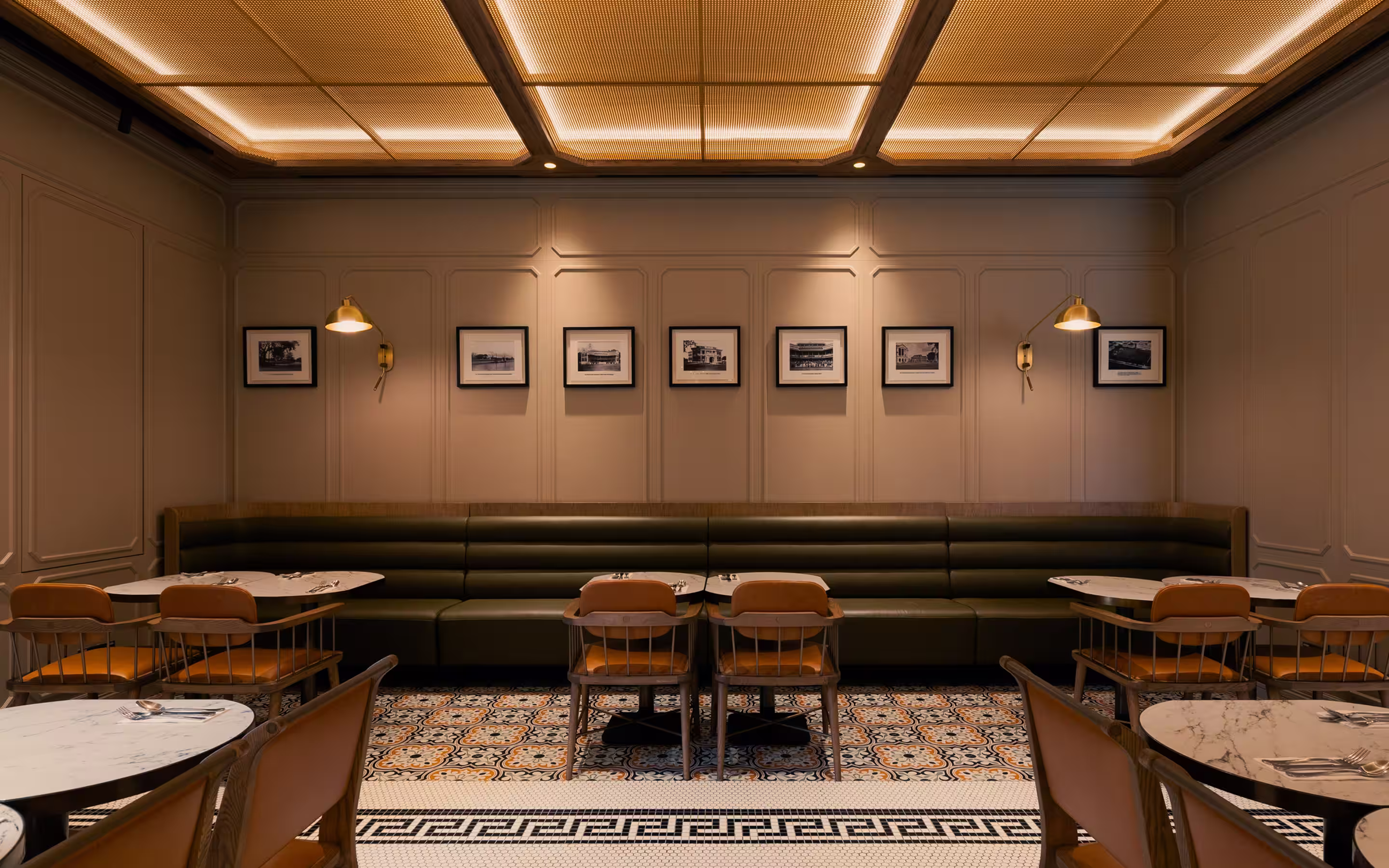 Elegant restaurant interior with green leather banquette, wooden chairs with orange cushions, marble tables, patterned tile floor, and framed black-and-white photos on beige walls.