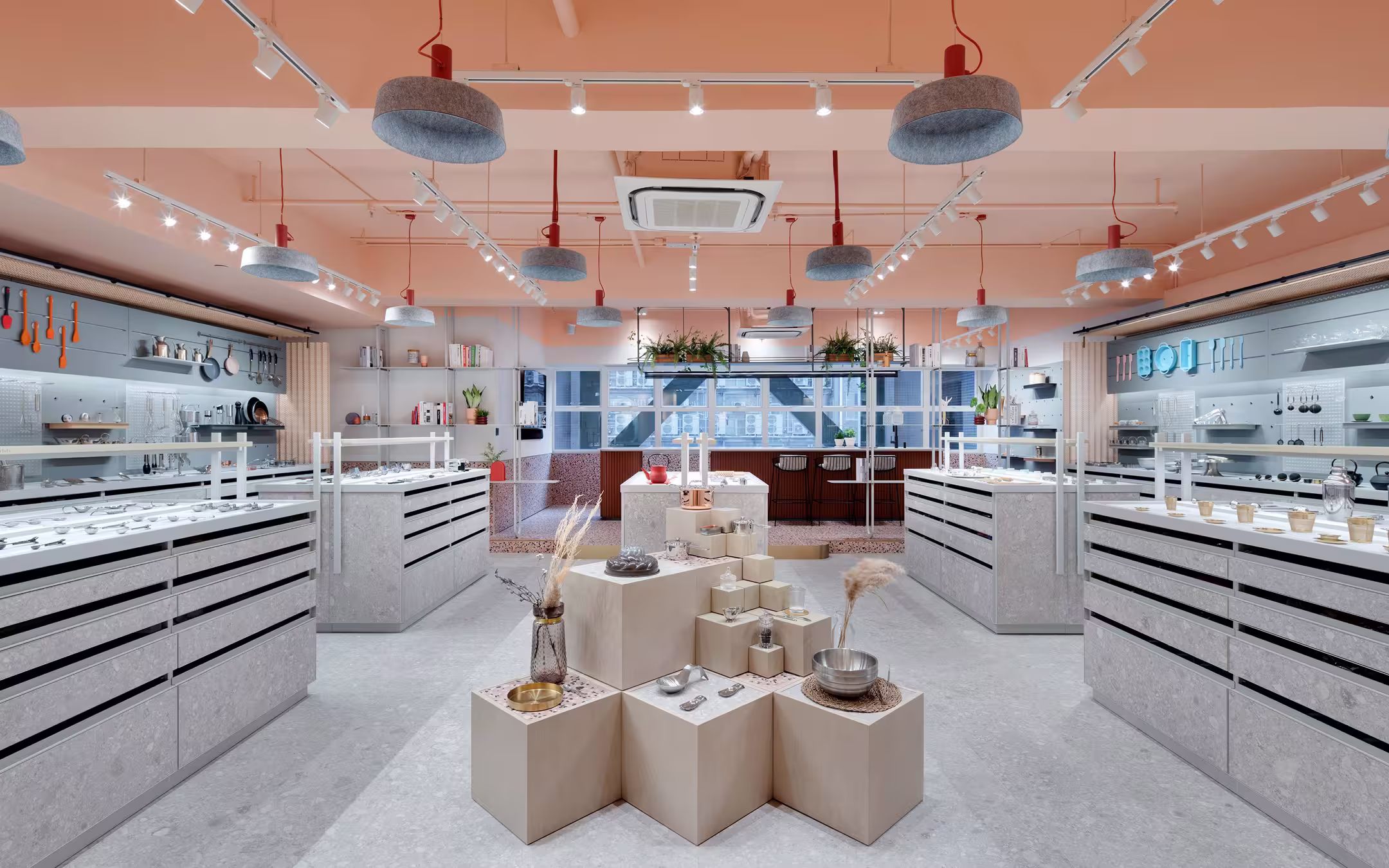 Modern store interior with minimalist display pedestals holding kitchen items, gray cabinets with utensils on walls, and large windows at the back.